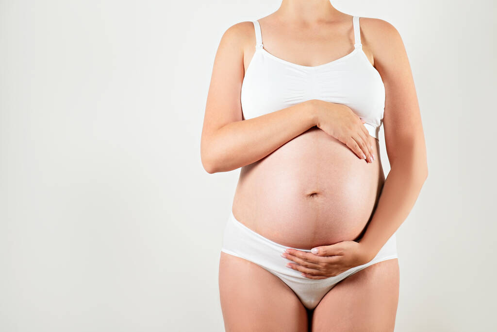 Close up of pregnant woman wearing supportive seamless maternity bra and maxi bottoms, arms on her belly. Female hands wrapped around big bare tummy. Child expectancy concept. Background, copy space - Photo, Image