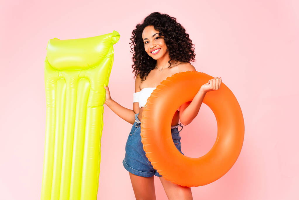 cheerful african american girl holding swim ring and inflatable mattress on pink  - Photo, Image