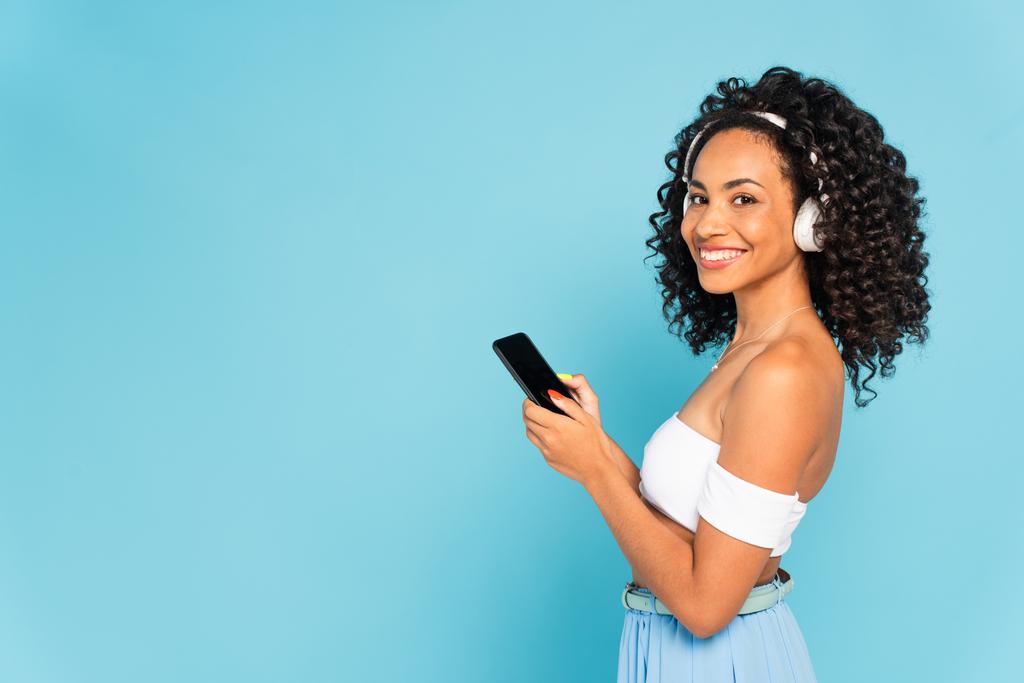 陽気なアフリカ系アメリカ人の女の子がワイヤレスヘッドフォンで音楽を聞いて、青い画面で隔離された空白の画面を持つスマートフォンを保持 - 写真・画像