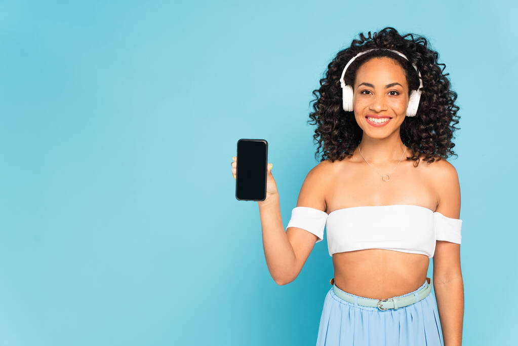feliz mulher africana americana ouvir música em fones de ouvido sem fio e segurando smartphone com tela em branco isolado no azul
 - Foto, Imagem