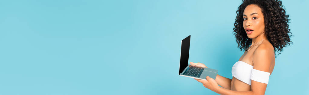 panoramic concept of surprised and curly african american woman holding laptop isolated on blue - Photo, Image