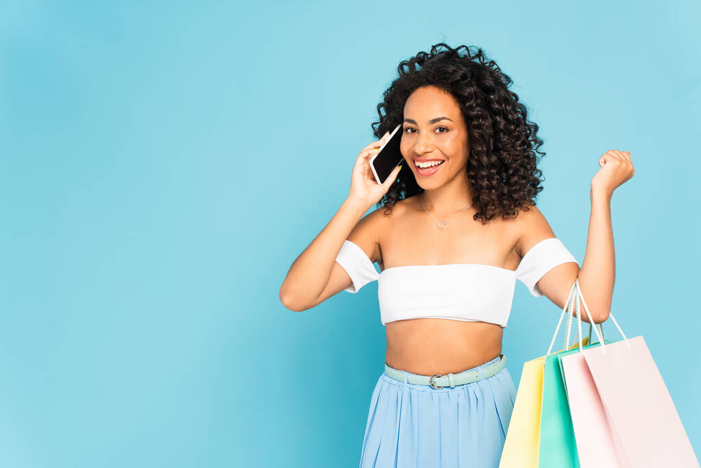 vidám afro-amerikai lány beszél okostelefonon, és gazdaság bevásárló táskák elszigetelt kék - Fotó, kép