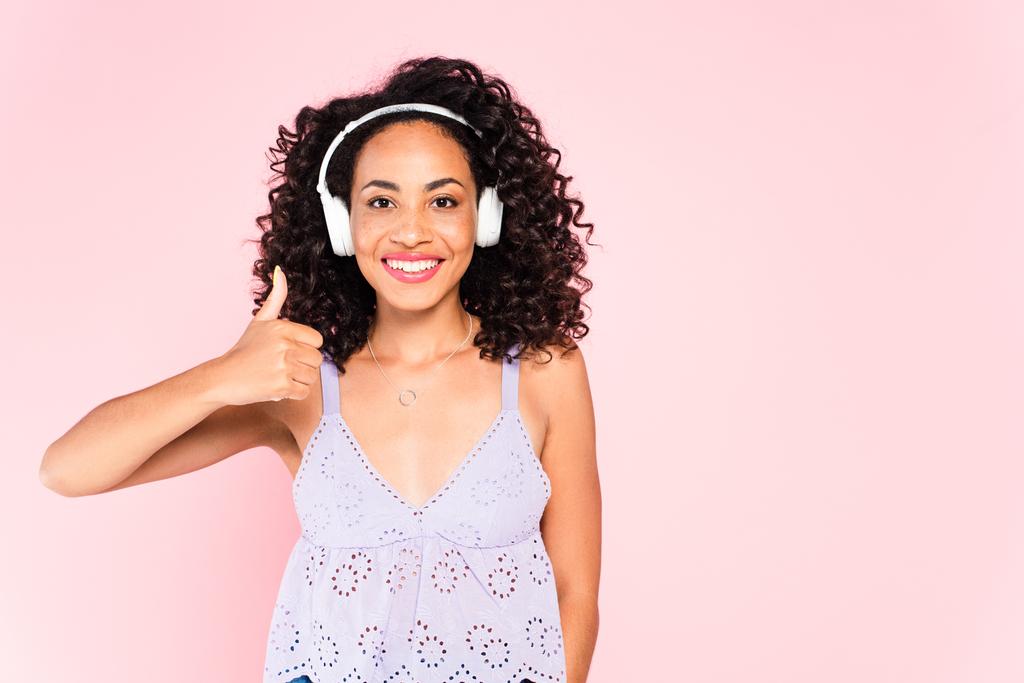 chica afroamericana feliz en auriculares inalámbricos escuchando música y mostrando el pulgar hacia arriba aislado en rosa
  - Foto, imagen