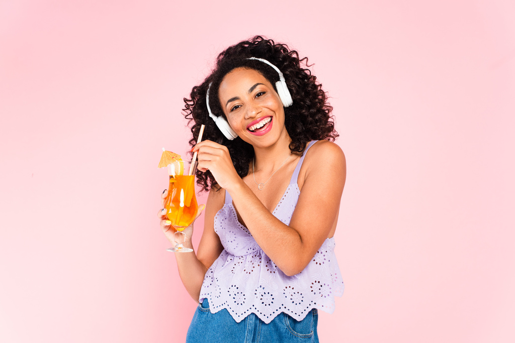 陽気なアフリカ系アメリカ人の女の子無線ヘッドフォンで音楽を聴きピンクに隔離されたカクテルを保持  - 写真・画像