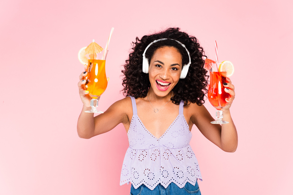 fröhliche afrikanisch-amerikanische Mädchen in drahtlosen Kopfhörern Musik hören und halten Cocktails isoliert auf rosa  - Foto, Bild