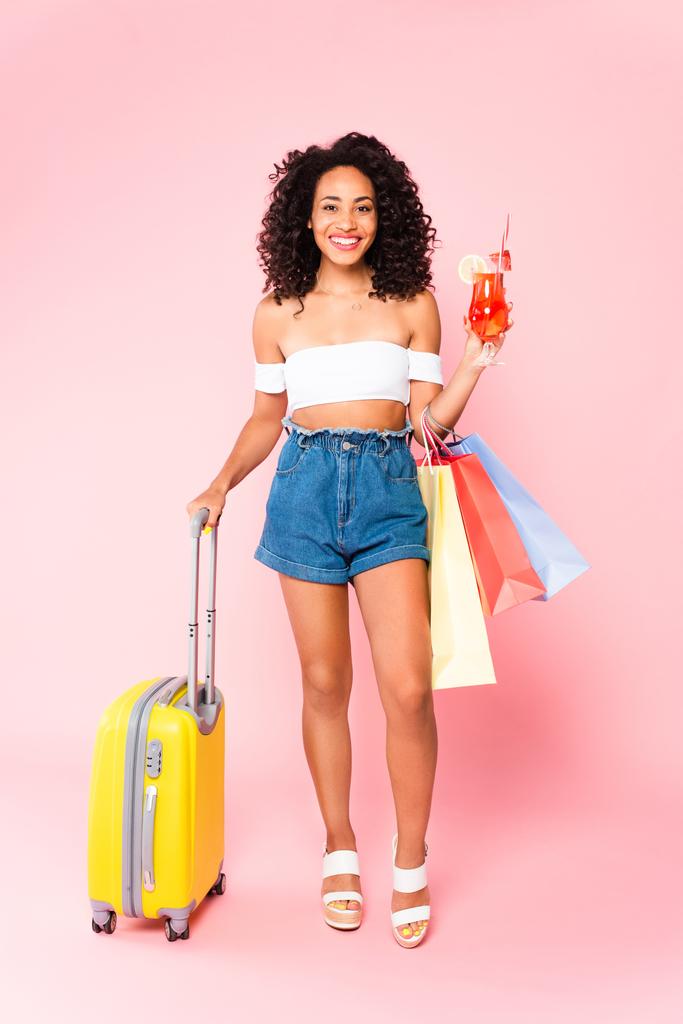 Gülümseyen Afro-Amerikalı kadın elinde kokteyl, alışveriş torbaları ve elinde bavulla pembenin üzerinde duruyor.  - Fotoğraf, Görsel
