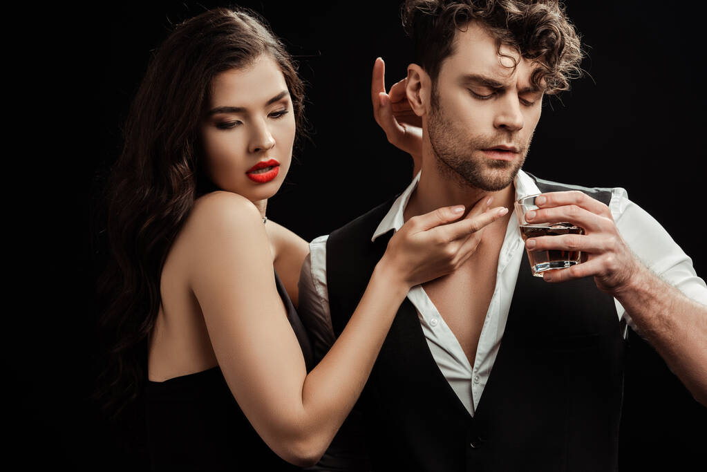 Beautiful woman with red lips touching boyfriend with glass of whiskey isolated on black  - Photo, Image