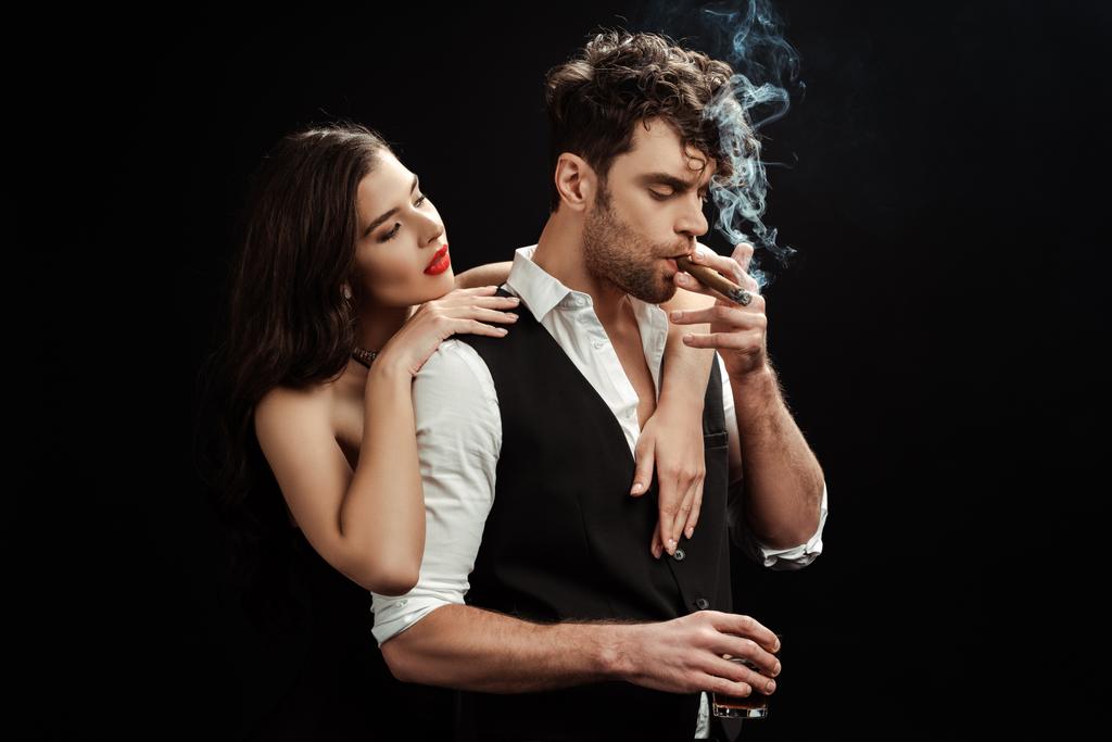 Beautiful girl embracing boyfriend with glass of whiskey and cigar isolated on black  - Photo, Image