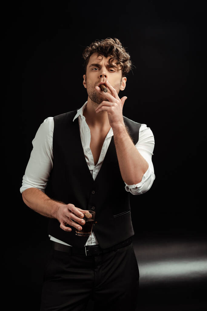 Handsome man looking at camera while smoking cigar and holding glass of whiskey on black background - Photo, Image