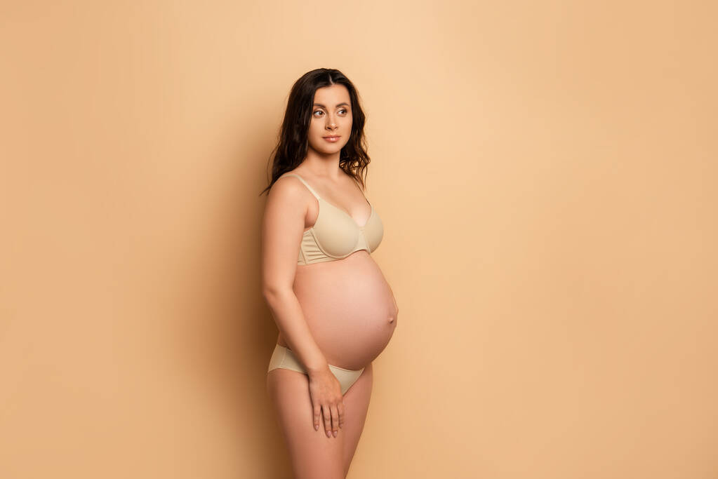 přemýšlivá těhotná žena ve spodním prádle dívá pryč, zatímco stojí na béžové - Fotografie, Obrázek