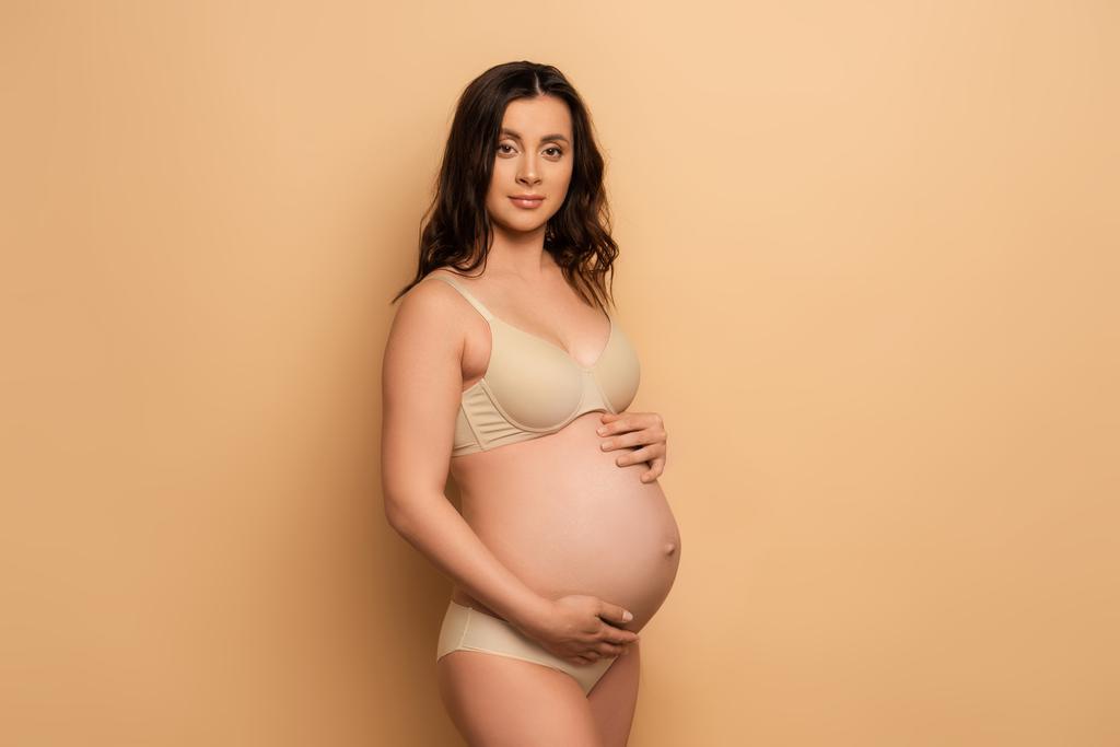 jovem mulher grávida em lingerie olhando para a câmera ao tocar barriga no bege
 - Foto, Imagem