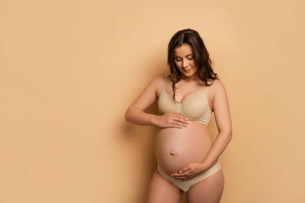 joven embarazada en ropa interior tocando la barriga en beige - Foto, imagen