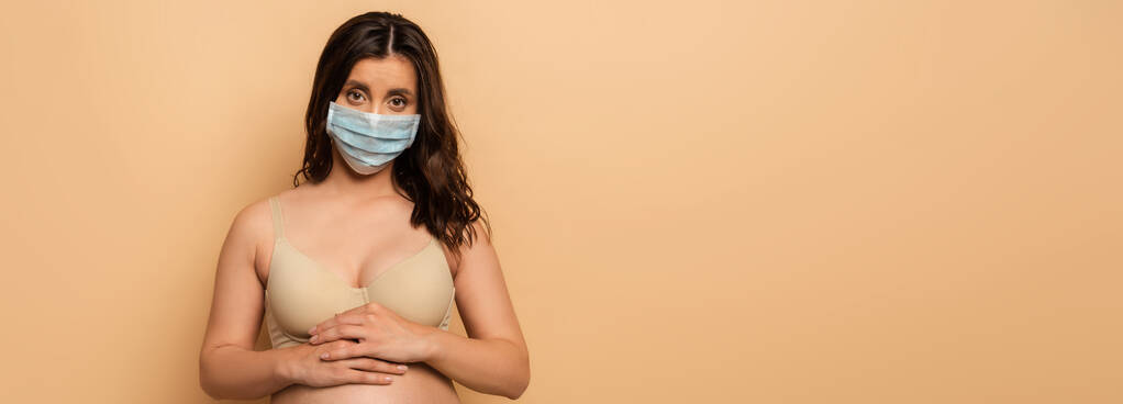 imagem horizontal de morena, mulher grávida em máscara protetora tocando barriga no bege
 - Foto, Imagem