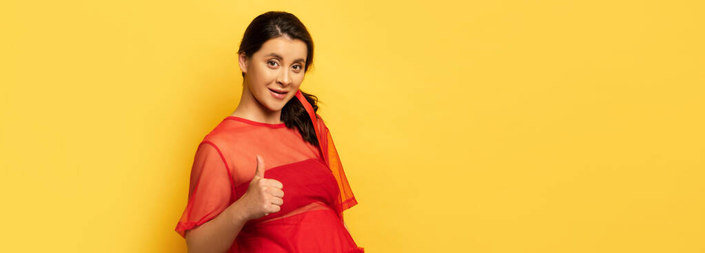 panoramatický koncept těhotné ženy v červené tunice ukazující palec nahoru při pohledu na fotoaparát na žluté - Fotografie, Obrázek