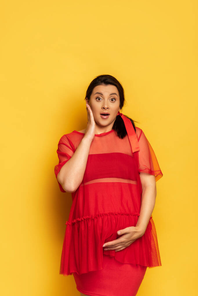 excited pregnant woman in red tunic touching tummy and face while looking at camera on yellow - Photo, Image