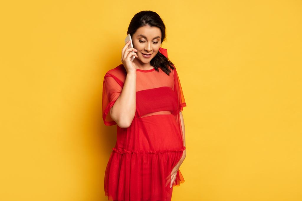 mladá těhotná žena v červené tunice dotýkající se břicha, zatímco mluví na smartphone na žluté - Fotografie, Obrázek