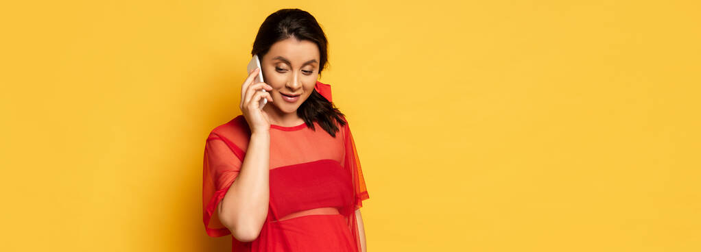 Kırmızı tunik giymiş hamile bir kadının sarı üzerine akıllı telefondan konuşmasının panoramik görüntüsü. - Fotoğraf, Görsel