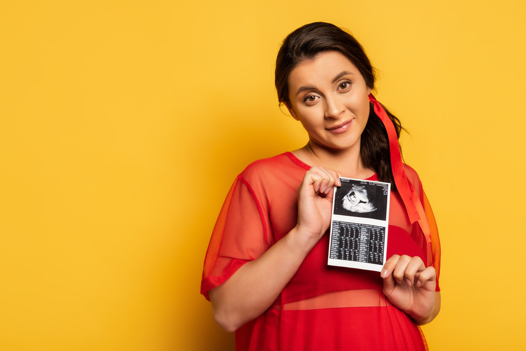 молодая беременная женщина в красном костюме проводит ультразвуковое сканирование, глядя в камеру на желтом - Фото, изображение