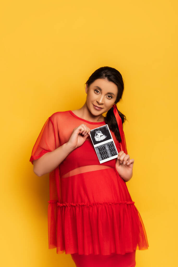 jonge zwangere vrouw in rode tuniek tonen echografie scan terwijl kijken naar camera op geel - Foto, afbeelding