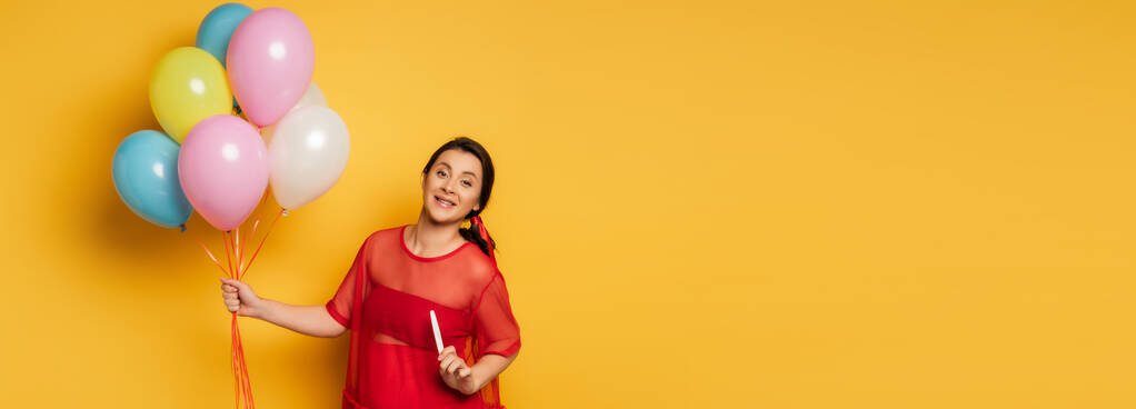 horizontales Bild einer schwangeren Frau im roten Outfit, die festliche Luftballons hält, während sie Schwangerschaftstest auf gelb zeigt - Foto, Bild