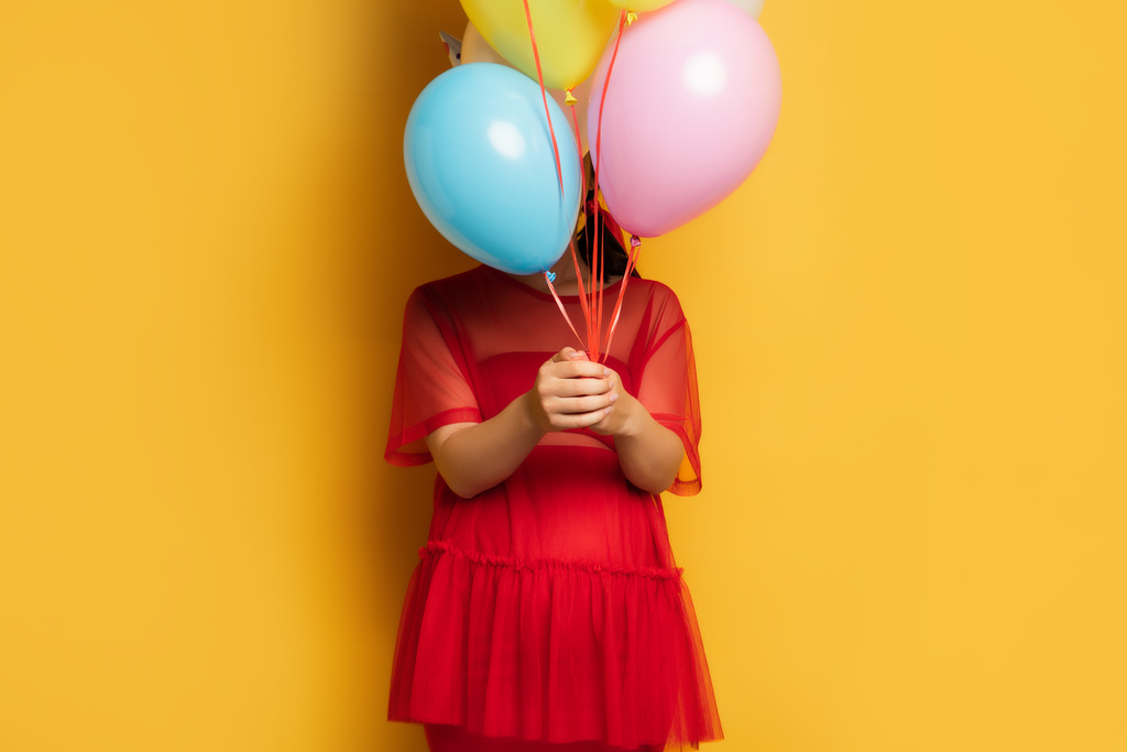 νεαρή έγκυος γυναίκα σε κόκκινο στολή σκίαση πρόσωπο με πολύχρωμα εορταστικά μπαλόνια στο κίτρινο - Φωτογραφία, εικόνα