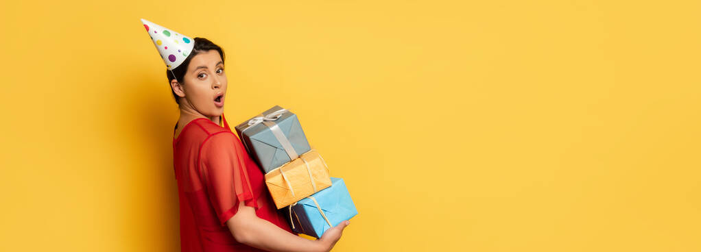 immagine orizzontale di donna incinta sorpresa in auto partito tenendo pila di regali mentre guardando la fotocamera su giallo - Foto, immagini