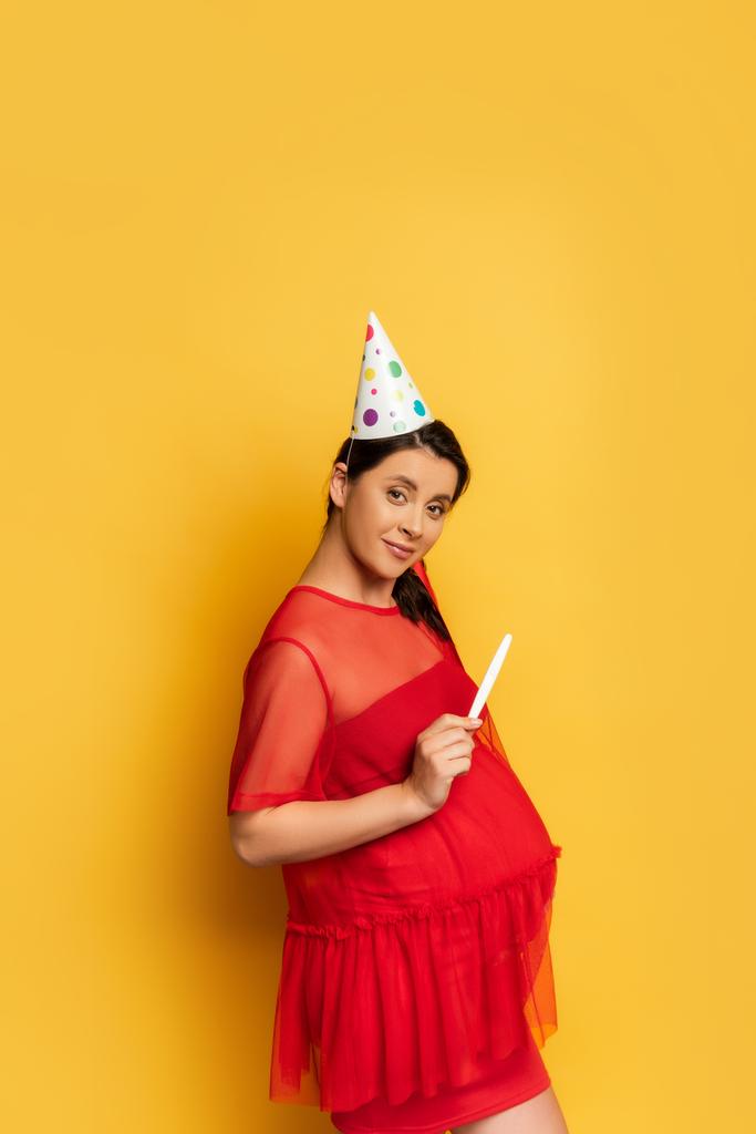 zwangere vrouw in feestmuts die zwangerschapstest toont terwijl ze naar de camera kijkt op geel - Foto, afbeelding