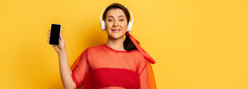 imagen horizontal de la mujer en auriculares inalámbricos que muestran el teléfono inteligente con pantalla en blanco en amarillo - Foto, imagen