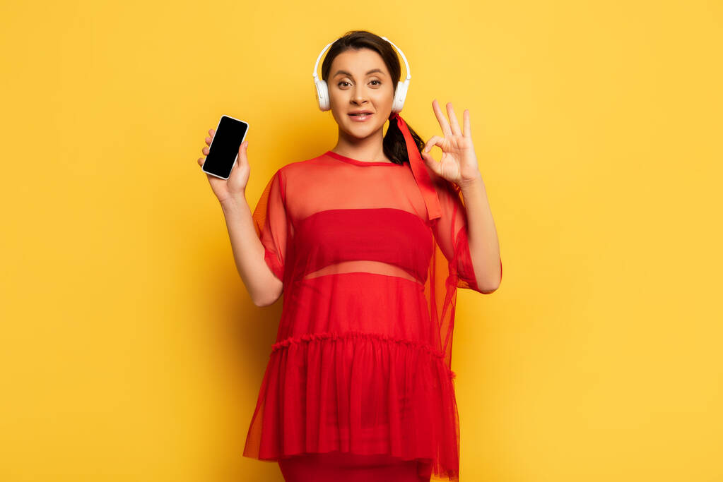 Schwangere mit drahtlosem Kopfhörer zeigt Smartphone und Okay-Geste auf Gelb - Foto, Bild