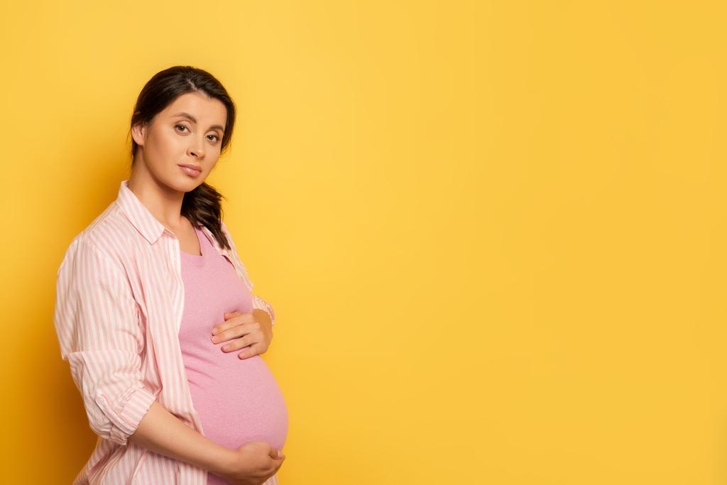 Genç hamile kadın sarıya dokunurken kameraya bakıyor. - Fotoğraf, Görsel