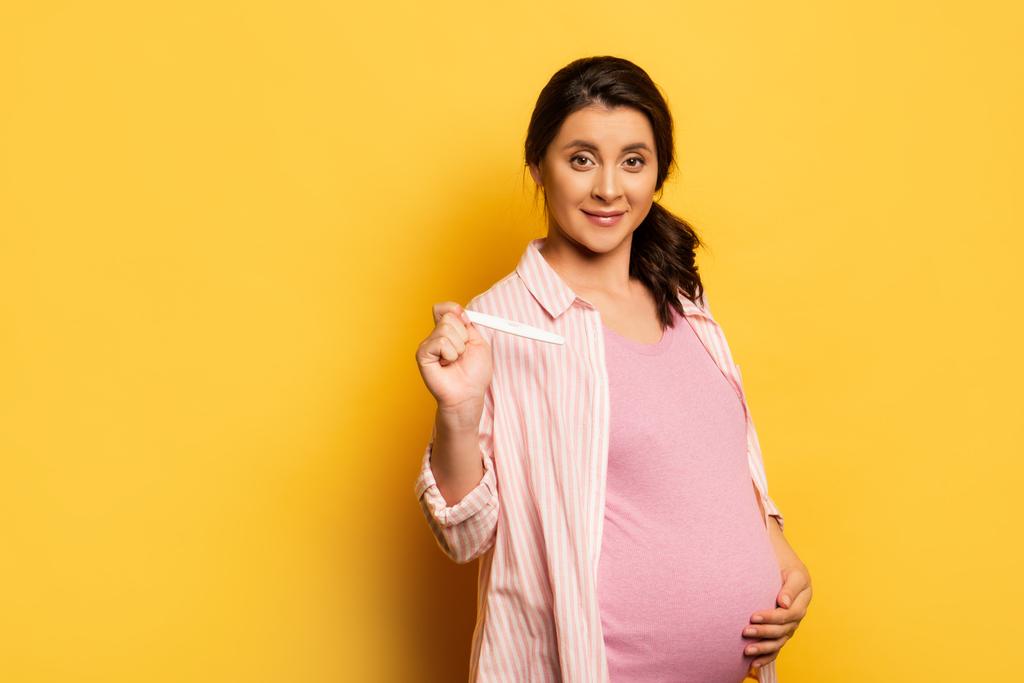 νεαρή γυναίκα που αγγίζει την κοιλιά και δείχνει τεστ εγκυμοσύνης στην κάμερα στο κίτρινο - Φωτογραφία, εικόνα