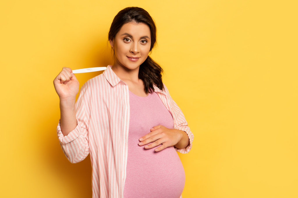 zwangere vrouw die de buik aanraakt en een zwangerschapstest op geel laat zien - Foto, afbeelding