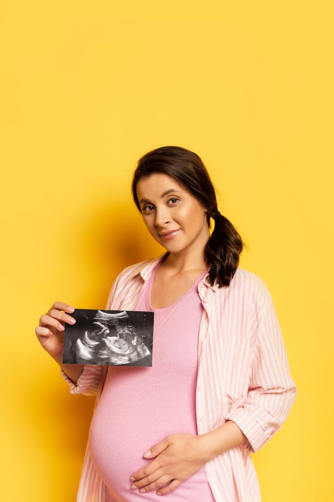 έγκυος γυναίκα δείχνει υπερηχογράφημα, ενώ αγγίζετε την κοιλιά σε κίτρινο   - Φωτογραφία, εικόνα