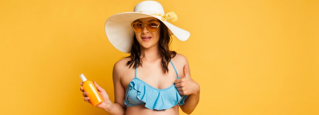 young woman in sunglasses and straw hat holding sunscreen and showing thumb up on yellow, horizontal image - Photo, Image