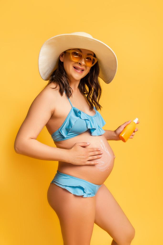 terhes nő alkalmaz naptejet a hasán, miközben a sárga kamerát nézi - Fotó, kép