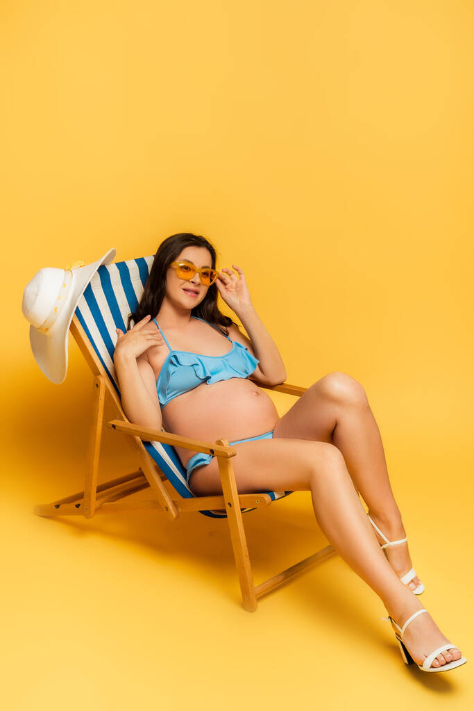 έγκυος γυναίκα με μπλε μαγιό αγγίζοντας γυαλιά ηλίου, ενώ κάθεται σε ξαπλώστρα στο κίτρινο - Φωτογραφία, εικόνα