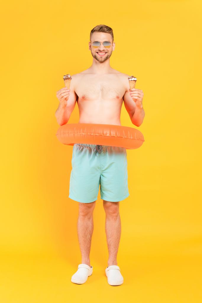 Mutlu ve üstsüz güneş gözlüklü, yüzme yüzüklü, elinde sarı külahta dondurma tutan bir adam. - Fotoğraf, Görsel