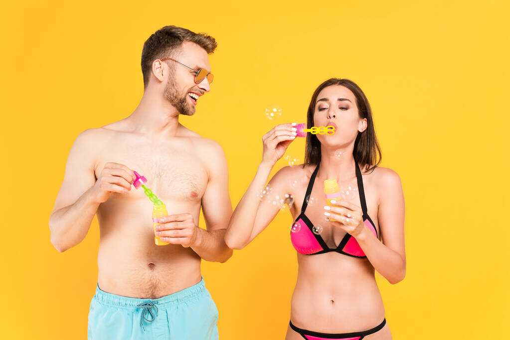 glücklicher und hemdloser Mann mit Sonnenbrille, der Mädchen im Badeanzug beim Blasen von Seifenblasen ansieht, isoliert auf gelb  - Foto, Bild