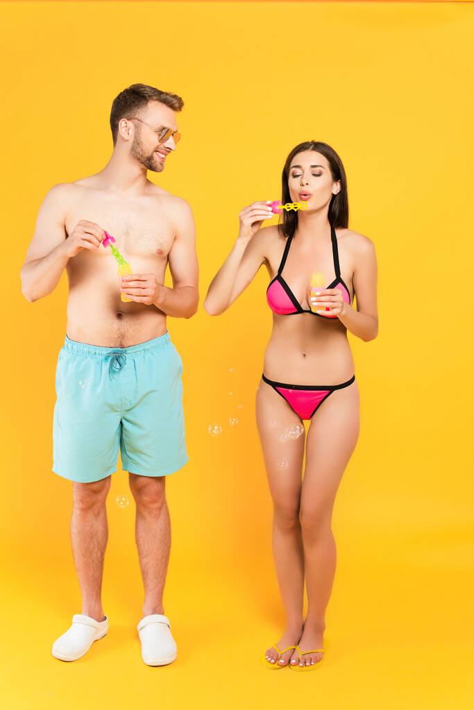 glücklicher und hemdloser Mann mit Sonnenbrille, der Mädchen im Badeanzug ansieht, die Seifenblasen auf gelb pusten  - Foto, Bild