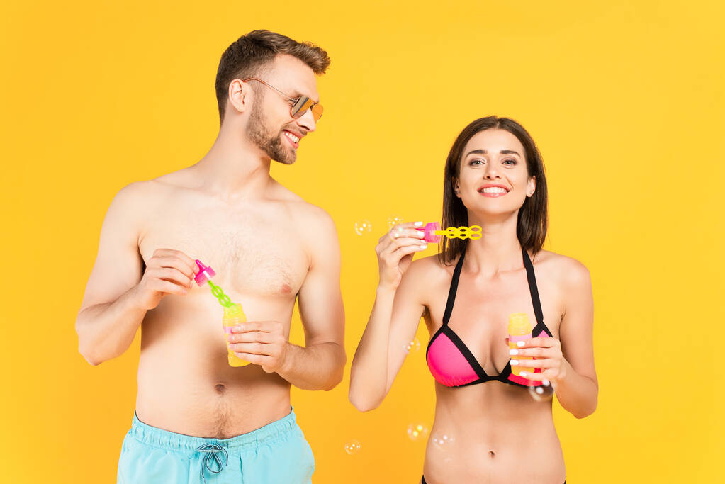 hombre alegre y sin camisa en gafas de sol mirando a la chica en traje de baño cerca de burbujas de jabón aislado en amarillo  - Foto, Imagen