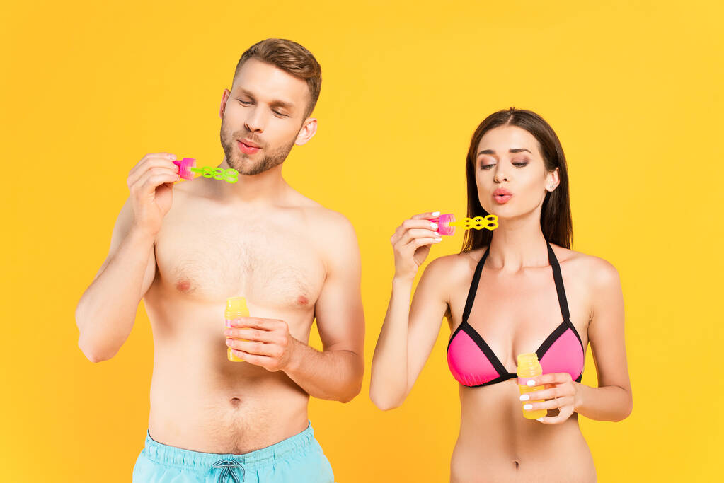 hombre sin camisa y atractiva chica en traje de baño soplando burbujas de jabón aislado en amarillo  - Foto, Imagen