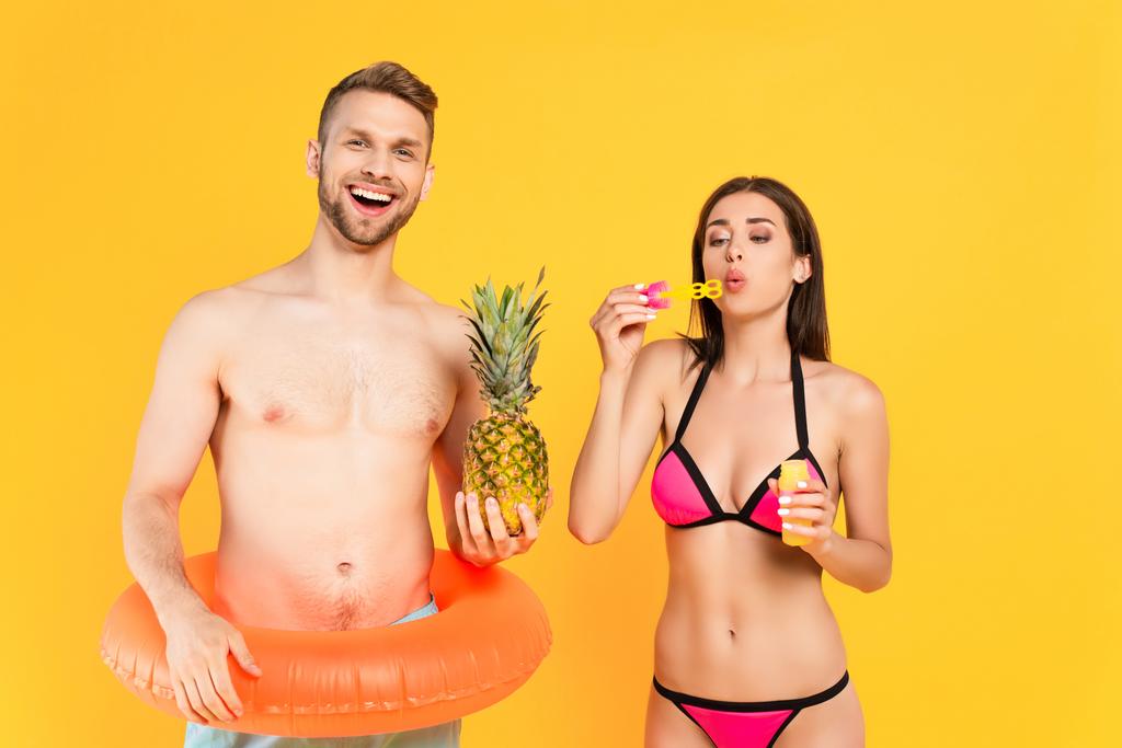 cheerful and shirtless man with swim ring holding pineapple near girl in swimsuit blowing soap bubbles isolated on yellow  - Photo, Image