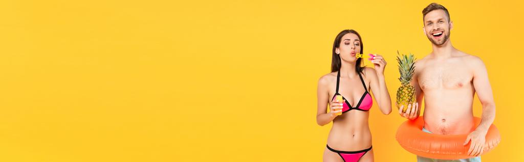 intestazione del sito web di uomo allegro con anello di nuoto in possesso di ananas vicino alla ragazza soffiando bolle di sapone isolato su giallo  - Foto, immagini