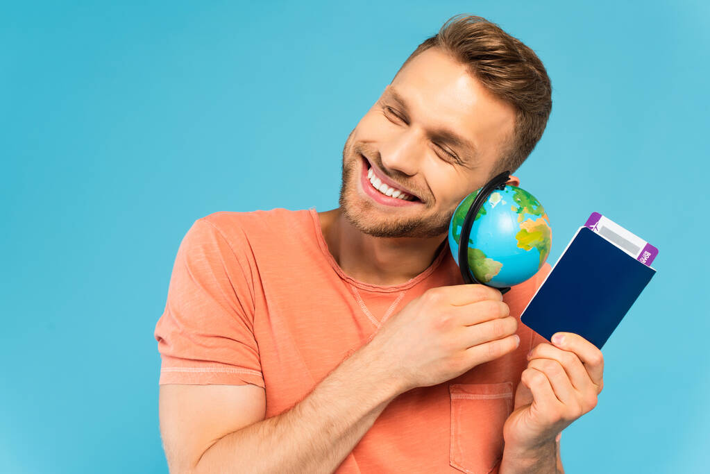gelukkig bebaarde man met gesloten ogen met paspoort en bol geïsoleerd op blauw - Foto, afbeelding