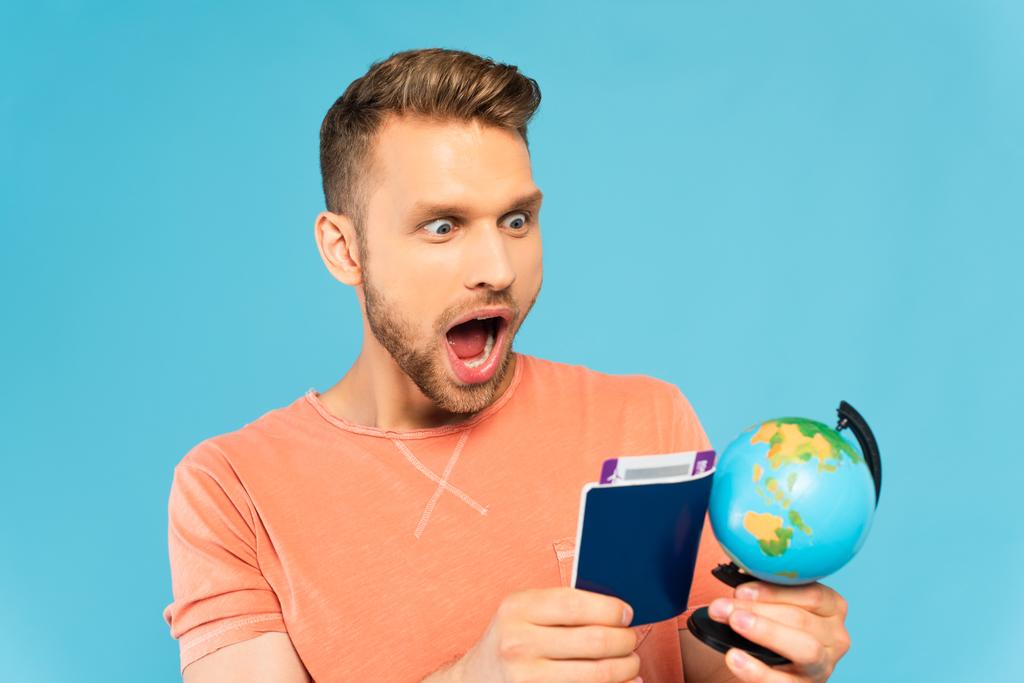 homme barbu choqué tenant passeport et regardant globe isolé sur bleu - Photo, image