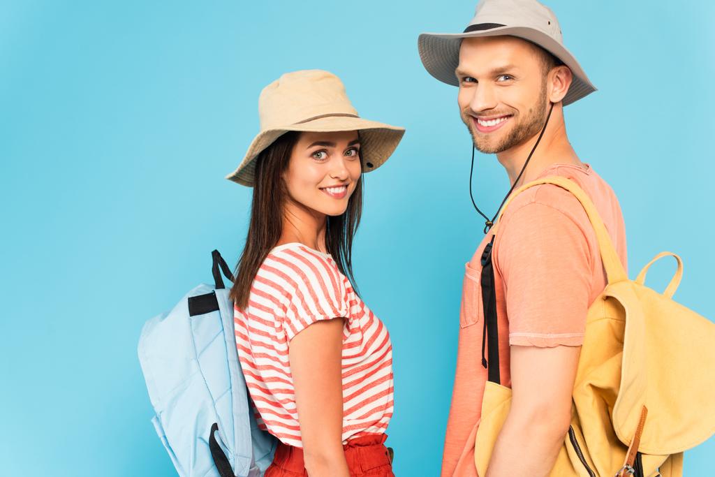 ευτυχισμένο ζευγάρι σε καπέλα με σακίδια κοιτάζοντας κάμερα απομονωμένη σε μπλε - Φωτογραφία, εικόνα