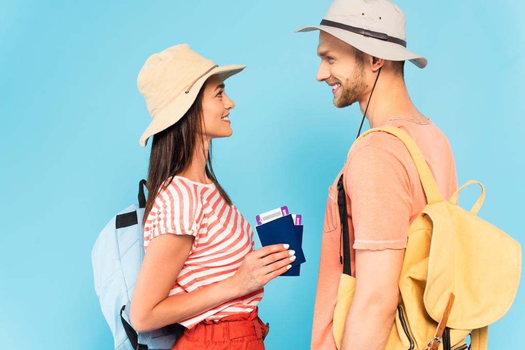 Şapkalı, elinde pasaportu olan mutlu bir kadının, mavi renkli izole edilmiş neşeli erkek arkadaşının yan görüntüsü. - Fotoğraf, Görsel