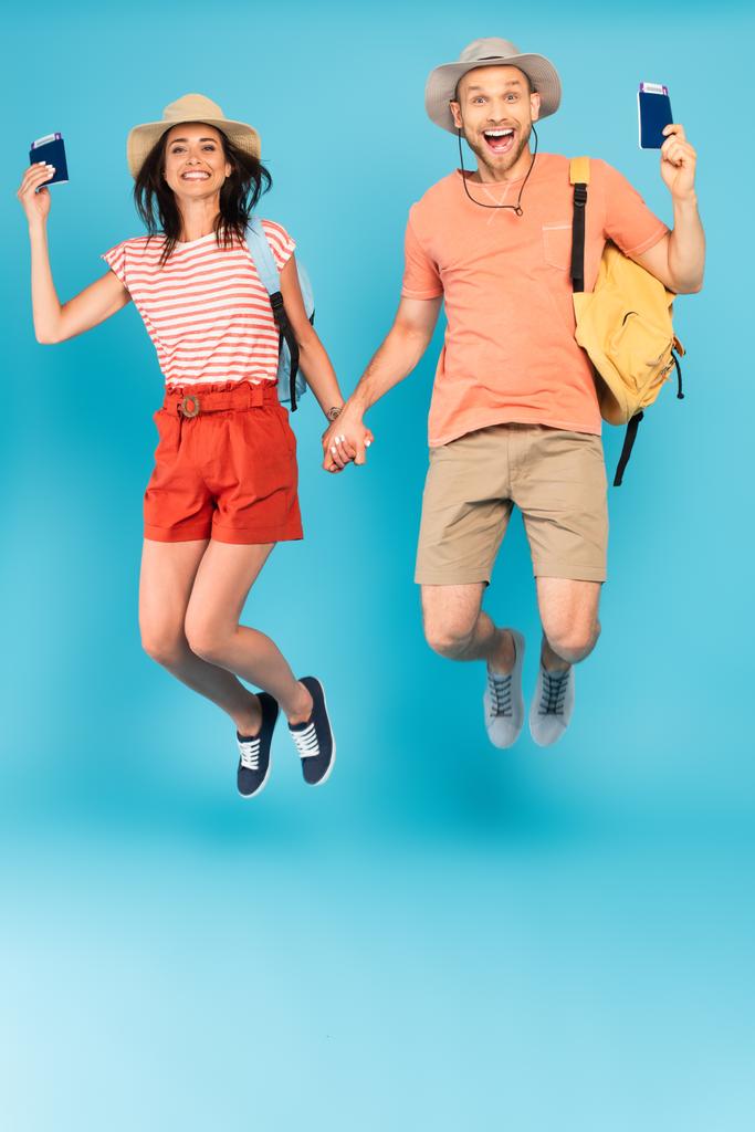 heureux couple chapeaux tenant la main et passeports tout en sautant sur bleu - Photo, image