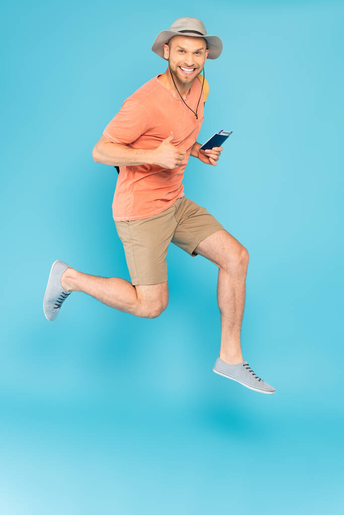 Χαρούμενος άνδρας με καπέλο κρατώντας διαβατήριο, πηδώντας και δείχνοντας τον αντίχειρα πάνω στο μπλε - Φωτογραφία, εικόνα