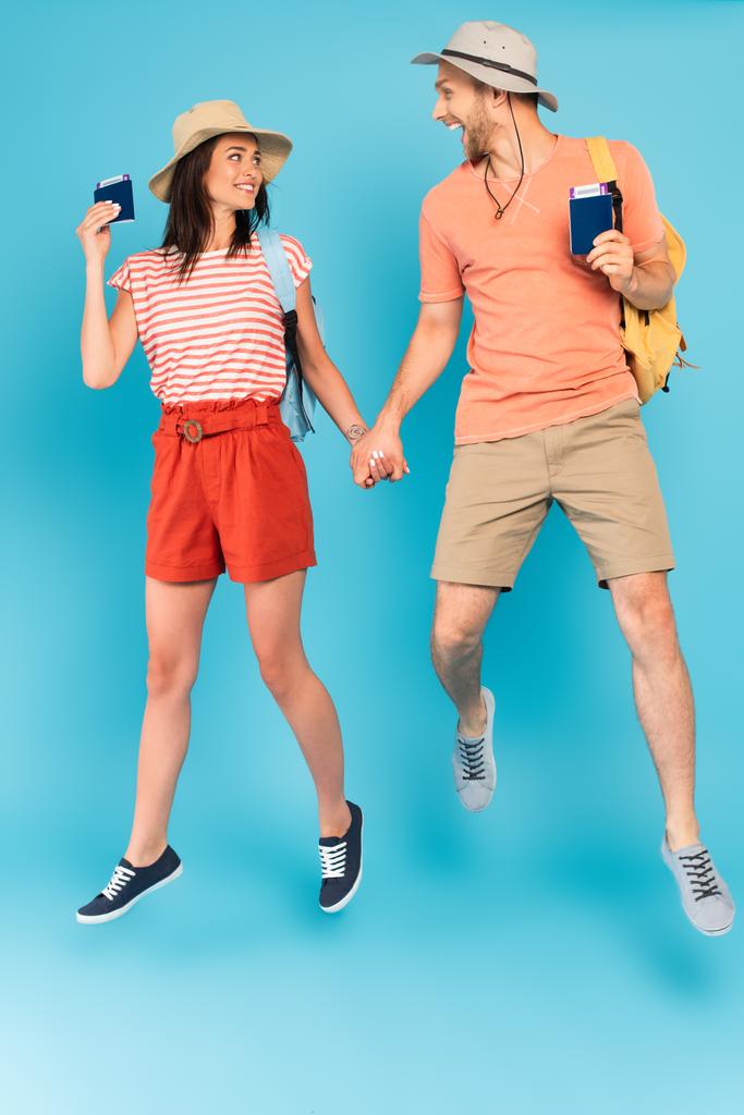 fröhliches Paar mit Hüten, die Hände und Pässe halten, während sie auf blau springen - Foto, Bild